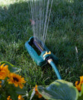 TurboTek Oscillating Sprinkler With 16 Precision Nozzles watering lawn surrounded by yellow flowers, flexible tubing visible.