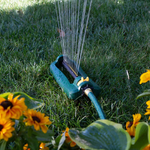 TurboTek Oscillating Sprinkler With 16 Precision Nozzles watering lawn surrounded by yellow flowers, flexible tubing visible.