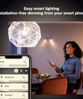 Woman using phone to control Dimmable White BR30 Smart Bulbs Starter Kit with Hub, 2 LED 60W equivalents, in a living room setting