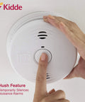 A hand pressing the hush button on a Dual Hazard Smoke And Carbon Monoxide Detector With Voice Warning Hardwired Setup And Ten Year Sealed Lithium Battery Backup Interconnectable Network Built Tough For Zero Drama Family Safety White.