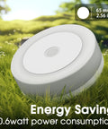 Close-up of white round LED night light with dimensions and energy saving features displayed in grassy outdoor setting.