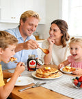 Family enjoying breakfast with pancakes, fresh fruit, and maple syrup served from a jar, in a bright kitchen setting.