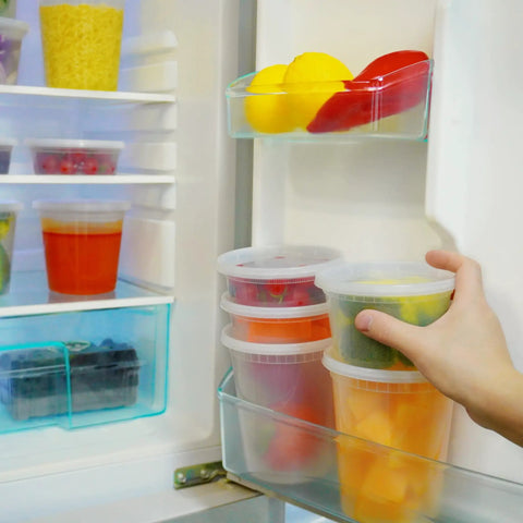 Hand placing colorful produce in 50 Pack 16 Oz Clear Plastic Deli Containers With Lids For Grab And Go Meal Prep in fridge door.