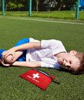 Red mini first aid kit pouch with zipper and white cross symbol on grass beside a boy in a white shirt.