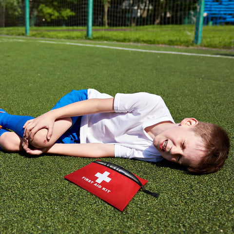 Red mini first aid kit pouch with zipper and white cross symbol on grass beside a boy in a white shirt.