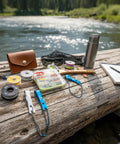 Stainless Steel Fishing Line Nipper Cutter with Keychain for fly fishing on a riverside log, with tools and a view of water.
