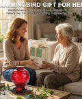 Two women exchanging a hummingbird feeder gift indoors, with a red Outdoor Hummingbird Feeder 25 Oz With Perch And Five Flower Ports in foreground.