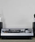 Crosley C100A Silver Belt Drive Turntable With Aluminum Tonearm Pitch Control For 33 And 45 Rpm Vinyl Records beside speakers and plants.