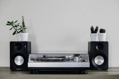 Crosley C100A Silver Belt Drive Turntable With Aluminum Tonearm Pitch Control For 33 And 45 Rpm Vinyl Records beside speakers and plants.