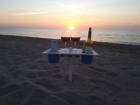 Portable Beach Table With Two Phone Slots, Lightweight Yet Sturdy And Durable on sand, holding wine and phone, sunset view.
