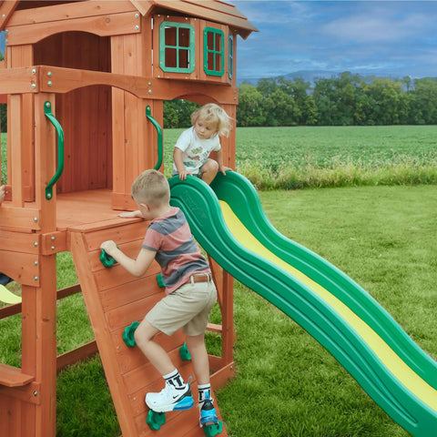 Children playing on Atlantis Cedar Wood Swing Set With Wave Slide And Raised Clubhouse Featuring Rock Wall in backyard.
