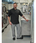 Badass Short Sleeve Chef Jacket In Sharp Black Features Double Breasted Front And Full Mesh Back For Sweet Kitchen Airflow Built From Tough Poly Cotton Twill That Fights Wrinkles And Holds Up Daily In Comfortable 2XL worn by a smiling chef in a kitchen.