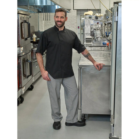Badass Short Sleeve Chef Jacket In Sharp Black Features Double Breasted Front And Full Mesh Back For Sweet Kitchen Airflow Built From Tough Poly Cotton Twill That Fights Wrinkles And Holds Up Daily In Comfortable 2XL worn by a smiling chef in a kitchen.