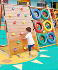 Child climbing on a wooden playground climbing wall with colorful handholds, next to a tire obstacle, in an outdoor play area.