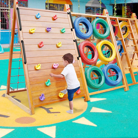 Child climbing on a wooden playground climbing wall with colorful handholds, next to a tire obstacle, in an outdoor play area.