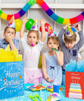 Children celebrating with Birthday Gift Bag Set With Handle And Tissue Paper, 12 Pack Assorted Sizes And Designs visible on table.