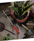 Top view of lightweight garden hand hoe with non-slip handle on soil, alongside a potted plant, shovel, and watering can.