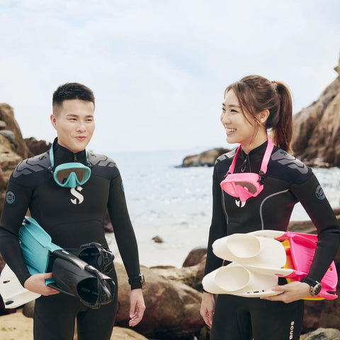 Two divers on rocky beach with wetsuits, snorkeling gear, and dive fins.