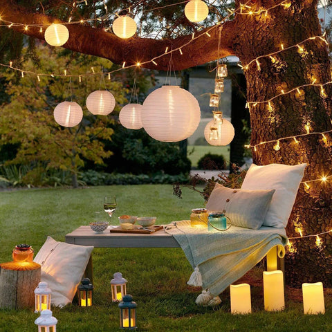 Outdoor scene with 12 Pack White LED Paper Lanterns in various sizes hanging from a tree, creating a cozy evening atmosphere.