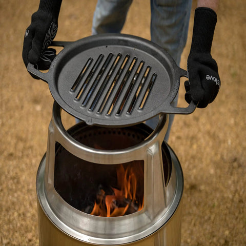Person holding a cast iron grill pan above a stainless steel fire pit outdoors.