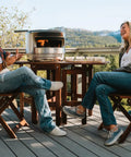 Pi Pizza Oven With Wood Or Gas Burner For Backyard Pizza Nights on wooden table with two people enjoying outdoor view