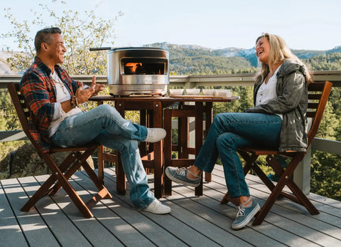 Pi Pizza Oven With Wood Or Gas Burner For Backyard Pizza Nights on wooden table with two people enjoying outdoor view