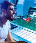 Technician inspecting printed circuit board being drilled with 10Pc PCB Drill Bit Set With One Eighth Inch Shank, Pro Grade Tungsten Carbide Micro Twist Bits For Printed Circuit Board, Jewelry, Plastic, Wood And Soft Metal, Sizes 0.3mm To 1.2mm, Includes Positioning Ring For Precise Drilling