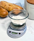 Glass bowl of flour on Primo Digital Food Scale For Precise Weighin, Portion Control, And Baking Prep, Easy Two Button Operation, Sealed Panel, Durable Chrome Finish, Compact Kitchen Scale For Grams And Ounces on countertop with bread.