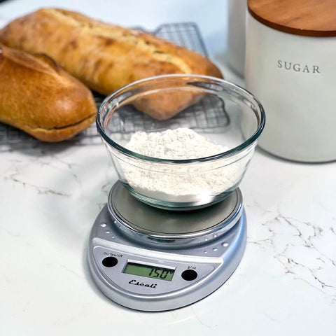 Glass bowl of flour on Primo Digital Food Scale For Precise Weighin, Portion Control, And Baking Prep, Easy Two Button Operation, Sealed Panel, Durable Chrome Finish, Compact Kitchen Scale For Grams And Ounces on countertop with bread.