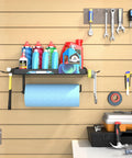 One Hand Tear Garage Paper Towel Holder With Shelf For Wall Mount 12 Inch Shop Paper Towel Dispenser Heavy Duty Iron Black Finish Super Handy Includes 2 Detachable Hooks For Garage And Tool Room, holding blue paper towel and bottles, installed on garage wall.