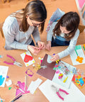 Two people crafting with the One Hole Punch With Soft Grip Handle For Paper One Quarter Inch Hole Punch Pink, surrounded by colorful paper.