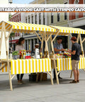 Collapsible Wooden Vendor Cart With Canopy Hella Spacious Market Stall Rack For Street Food And Outdoor Events Built Tough With Quick Snap Assembly Sweet Olive Green Finish Plus Ample Counter Space For Your Mobile Business Gear in a sunny street market.