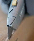Close-up of everyday utility knife with retractable blade cutting cardboard, silver non-slip handle visible.