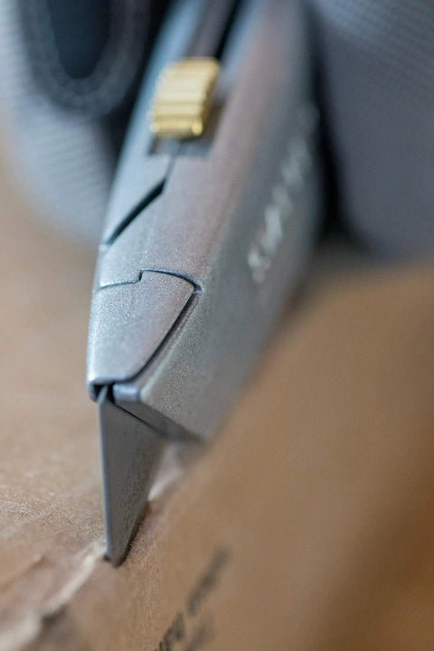Close-up of everyday utility knife with retractable blade cutting cardboard, silver non-slip handle visible.