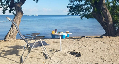 Portable Beach Table With Two Phone Slots, Lightweight Yet Sturdy And Durable, set up on sandy beach beside a chair with cups.