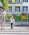 Woman and child crossing street beside a Slow Down Kids At Play Road Sign For Neighborhoods, Aluminum Weatherproof Safety Plaque