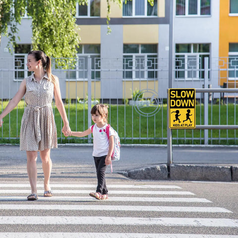 Woman and child crossing street beside a Slow Down Kids At Play Road Sign For Neighborhoods, Aluminum Weatherproof Safety Plaque