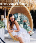 Woman sitting in hanging basket chair with floral cushion under a thatched roof in outdoor setting