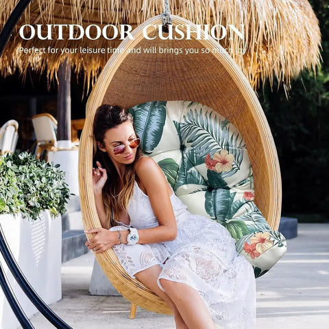 Woman sitting in hanging basket chair with floral cushion under a thatched roof in outdoor setting