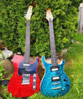 Two electric guitars with red and blue quilted wood finishes leaning against a green hedge outdoors.