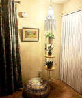 Macrame hanging shelf with plants in cozy corner, featuring intricate design against warm lit wall with framed art.
