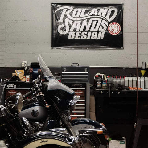 Motorcycle parked in garage with tool storage and banner labeled 'Roland Sands Design' on the wall.