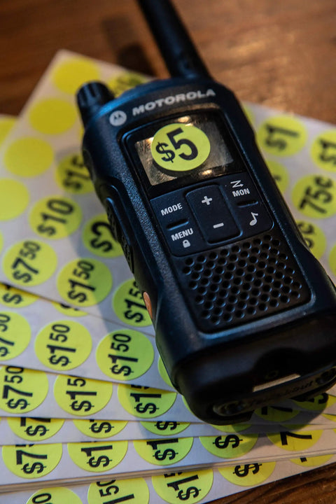 Close-up of a walkie-talkie placed on yellow $5 and $10 price stickers from Yard Sale EZ Kit Neon Yellow Signs 11 By 14 Inches With 600 Prepriced Stickers Price Label Sheets And Arrows For Directing Customers Includes 15 Writable Large Stickers And Repriced Sticker Pack For Fast Pricing.