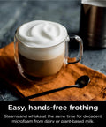 Glass mug of coffee with frothy milk next to spoon showcasing frothing feature of R-ES601 Luxe Café Premier Series 3-In-1 Espresso Machine
