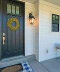 Front porch with white siding, a black door with wreath, lantern light, welcome mat, and bench with pillows