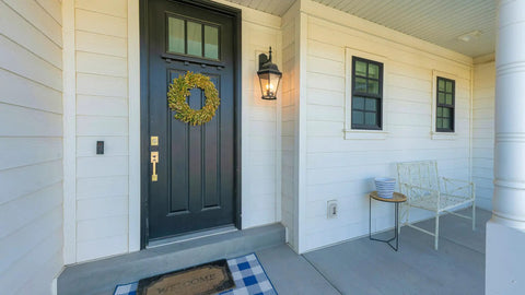 Front porch with white siding, a black door with wreath, lantern light, welcome mat, and bench with pillows