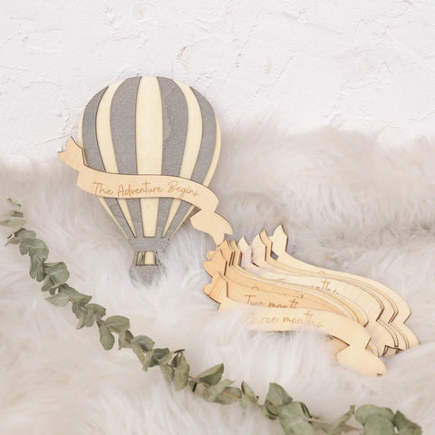 Wooden Hot Air Balloon Monthly Milestone Cards For Babies on a fluffy surface with leaves, featuring a striped balloon design.