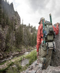 Angler hiking with Ultra Lightweight Fly Fishing Vest With 14 Pockets And Front Zip Storage Olive Green by a rocky river landscape.