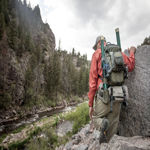 Angler hiking with Ultra Lightweight Fly Fishing Vest With 14 Pockets And Front Zip Storage Olive Green by a rocky river landscape.