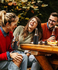 Group of friends enjoying drinks outdoors using Stainless Steel 12oz Vacuum Insulated Can And Bottle Cooler Keeps Ya Drinks Ice Cold With Double Wall Construction Copper Plated Chiller Metal Cooler For Cans And Bottles Black Finish on a picnic table in a forest setting.